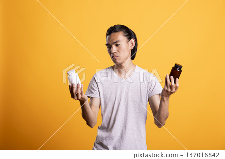 Man examines two different pill bottles, feeling unsure which medication to choose. Person compares white and brown containers with confused expression, showing decision making in personal health care Man examines two different pill bottles, feeling unsure which medication to choose. Person compares white and brown containers with confused expression, showing decision making in personal health care 137016842