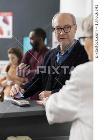 Senior white man tapping credit card to pay for medicine in modern pharmacy checkout. The scene captures everyday customer service and digital payment convenience in healthcare environment. 137016843