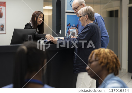 Professional hospital administrator assists pensioner patients with registration and appointment forms in modern clinic. Asian receptionist helps retired couple in filling medical forms for checkup. Professional hospital administrator assists pensioner patients with registration and appointment forms in modern clinic. Asian receptionist helps retired couple in filling medical forms for checkup. 137016854