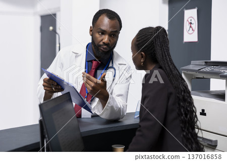 Black male doctor discussing patient information with administrator at reception desk. Two healthcare workers reviewing health insurance form on clipboard and confirming details for medical record. Black male doctor discussing patient information with administrator at reception desk. Two healthcare workers reviewing health insurance form on clipboard and confirming details for medical record. 137016858