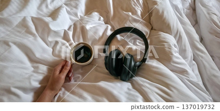 A woman's hand holds a cup of coffee and headphones lie nearby on light-colored bed linens. Morning routine. High quality photo A woman's hand holds a cup of coffee and headphones lie nearby on light-colored bed linens. Morning routine. High quality photo 137019732