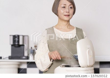 一位老婦人正在廚房裡把剛煮好的飯從電鍋裡倒進碗裡。 一位老婦人正在廚房裡把剛煮好的飯從電鍋裡倒進碗裡。 137020312