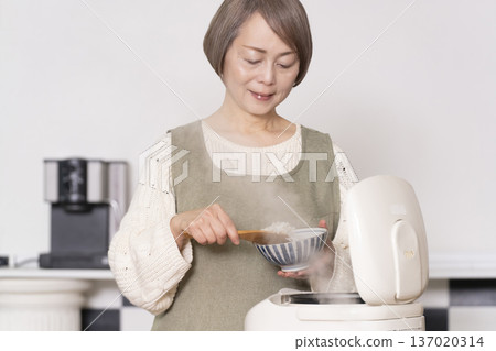 一位老婦人正在廚房裡把剛煮好的飯從電鍋裡倒進碗裡。 137020314
