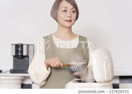 一位老婦人正在廚房裡把剛煮好的飯從電鍋裡倒進碗裡。 137020315