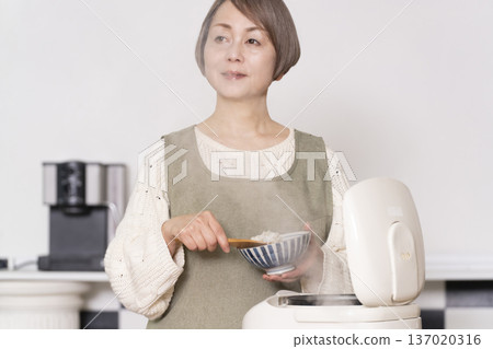 一位老婦人正在廚房裡把剛煮好的飯從電鍋裡倒進碗裡。 137020316