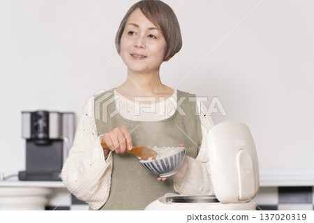 一位老婦人正在廚房裡把剛煮好的飯從電鍋裡倒進碗裡。 一位老婦人正在廚房裡把剛煮好的飯從電鍋裡倒進碗裡。 137020319