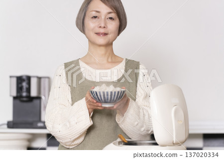 一位老婦人正在廚房裡把剛煮好的飯從電鍋裡倒進碗裡。 137020334