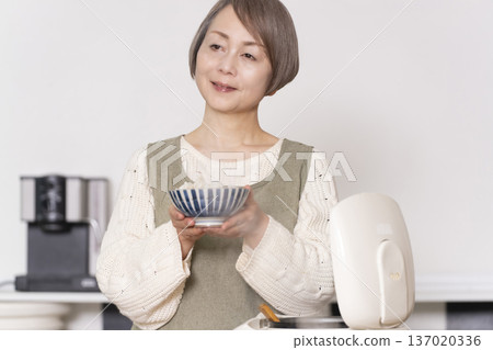 一位老婦人正在廚房裡把剛煮好的飯從電鍋裡倒進碗裡。 一位老婦人正在廚房裡把剛煮好的飯從電鍋裡倒進碗裡。 137020336