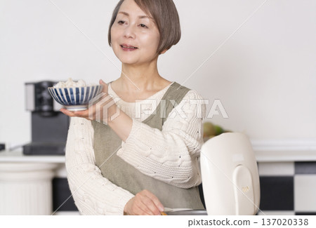 一位老婦人正在廚房裡把剛煮好的飯從電鍋裡倒進碗裡。 137020338