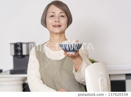 一位老婦人正在廚房裡把剛煮好的飯從電鍋裡倒進碗裡。 137020339