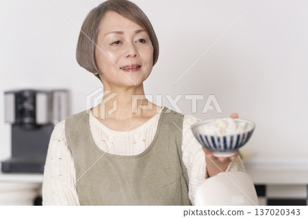 A senior woman pouring freshly cooked rice from a rice cooker into a bowl in the kitchen 137020343