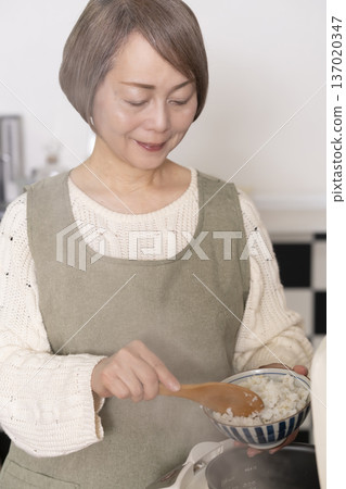 A senior woman pouring freshly cooked rice from a rice cooker into a bowl in the kitchen 137020347