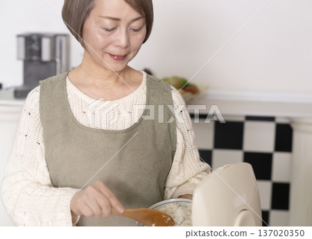 一位老婦人正在廚房裡把剛煮好的飯從電鍋裡倒進碗裡。 137020350