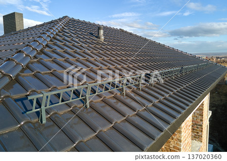 Closeup snow guard for safety in winter on house roof top covered with ceramic shingles. Tiled covering of building 137020360