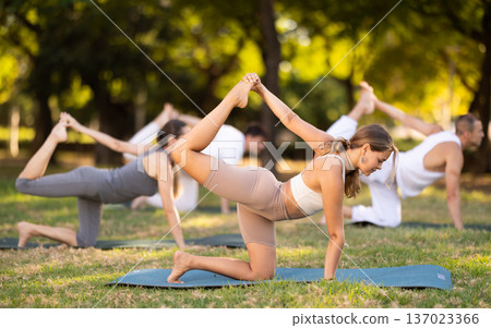 Young yogini performing Vyaghrasana asana during group yoga outdoors Young yogini performing Vyaghrasana asana during group yoga outdoors 137023366