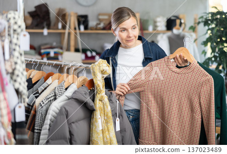 Fashionable european woman in casual clothes standing near clothes rack and choosing new dress for autumn-winter season in clothing boutique Fashionable european woman in casual clothes standing near clothes rack and choosing new dress for autumn-winter season in clothing boutique 137023489