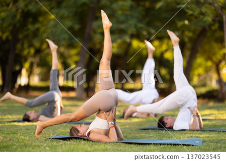 Athletic young girl demonstrating flexibility in modified Shoulder Stand Eka Pada Sarvangasana during group yoga training on green meadow in summer park Athletic young girl demonstrating flexibility in modified Shoulder Stand Eka Pada Sarvangasana during group yoga training on green meadow in summer park 137023545