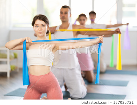 Happy athletic muscular young girl performing workout routine in gym with group of sporty people, kneeling on mat and doing exercise with pilates resistance bands 137023721