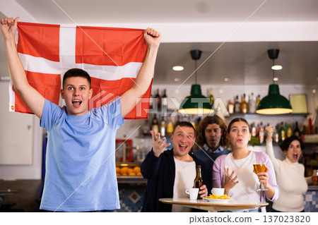 Screaming young adult sports fans rooting for favorite team and waving flag of Denmark while watching match together in pub 137023820
