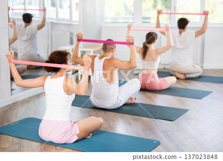 Back view of young girl with group of young adults maintaining physical health practicing pilates with resistance band sitting on gymnastics mat at fitness center Back view of young girl with group of young adults maintaining physical health practicing pilates with resistance band sitting on gymnastics mat at fitness center 137023848