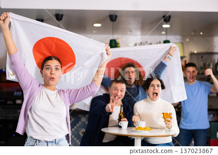 Company of emotive young adult friends rooting for favorite team and waving flag of Japan while watching match together in bar 137023867