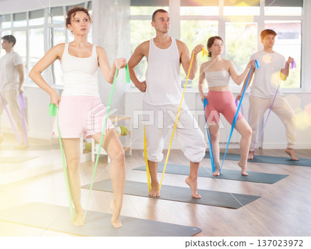 Positive woman with sporty women and men engaged in group class standing and perform exercise with resistance bands in pilates studio Positive woman with sporty women and men engaged in group class standing and perform exercise with resistance bands in pilates studio 137023972