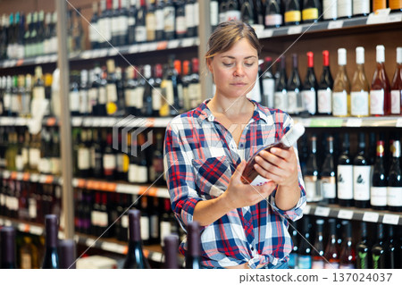 Young woman carefully reads the label on bottle while choosing wine in supermarket Young woman carefully reads the label on bottle while choosing wine in supermarket 137024037