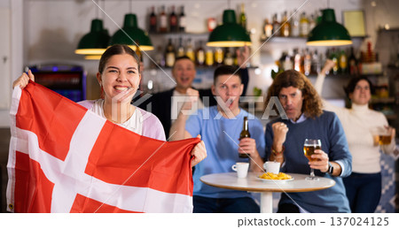 Company of young adult enthusiastic fans waving flag of Denmark and supporting national team with beer in pub 137024125