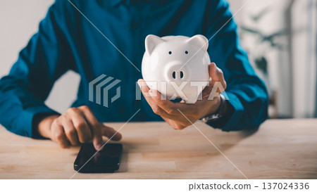 Fintech bank piggy money save mobile finance app financial phone savings, man holding white ceramic pig while using smartphone for digital payment at wooden office desk table 137024336