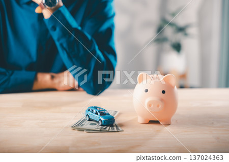 Man uses pink piggy bank and toy car on money to manage gdp savings insurance finance health budget and save for vehicle purchase while sitting at wooden home office table surface Man uses pink piggy bank and toy car on money to manage gdp savings insurance finance health budget and save for vehicle purchase while sitting at wooden home office table surface 137024363