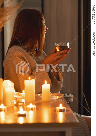 Woman in bathrobe enjoying hot tea illuminated by flickering candles, finding peace and tranquility. Wellness and spa evening Woman in bathrobe enjoying hot tea illuminated by flickering candles, finding peace and tranquility. Wellness and spa evening 137025666