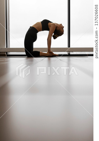 A woman performs the Ustrasana yoga pose, also known as Camel Pose, in a bright studio with large windows 137026608