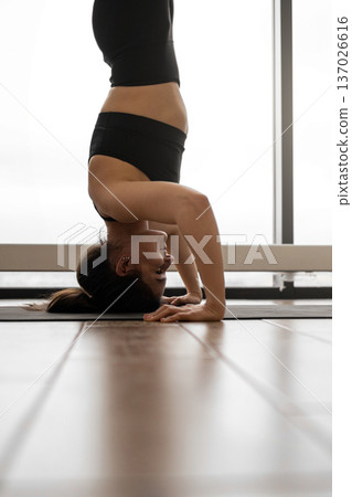 A woman in black athletic wear performs a headstand yoga pose on a mat in a bright room with wooden floors and large windows A woman in black athletic wear performs a headstand yoga pose on a mat in a bright room with wooden floors and large windows 137026616