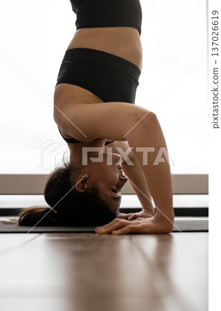A woman in black athletic wear performs a headstand on a yoga mat, demonstrating balance and strength A woman in black athletic wear performs a headstand on a yoga mat, demonstrating balance and strength 137026619