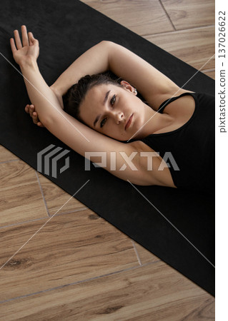 A young woman in athletic wear rests on a black yoga mat, her arms crossed above her head, looking thoughtfully towards the camera 137026622