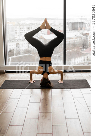 A woman in black activewear performs a headstand yoga pose on a mat in front of a large window overlooking a city skyline 137026643