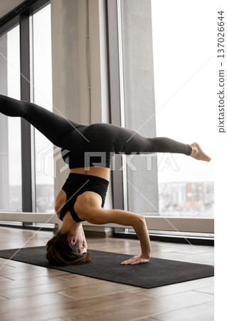 A woman in black athletic wear performs a challenging yoga headstand on a mat in a bright room with large windows A woman in black athletic wear performs a challenging yoga headstand on a mat in a bright room with large windows 137026644