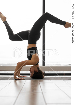 A woman in black activewear performs a challenging yoga headstand pose with her legs extended upwards against a bright window backdrop 137026652