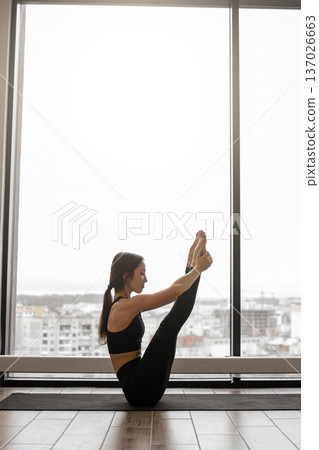 A woman in black activewear performs a yoga pose, balancing on her mat with legs extended towards the ceiling, framed by a large window overlooking a city skyline A woman in black activewear performs a yoga pose, balancing on her mat with legs extended towards the ceiling, framed by a large window overlooking a city skyline 137026663