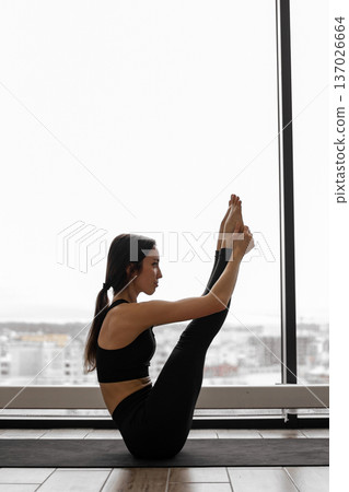 A woman in black activewear performs a yoga pose, balancing on her glutes with legs extended upwards, near a large window with a city view A woman in black activewear performs a yoga pose, balancing on her glutes with legs extended upwards, near a large window with a city view 137026664