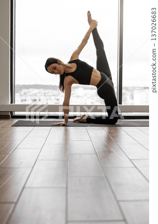 A woman in black activewear performs a challenging yoga pose on a mat in front of a large window with a city view 137026683