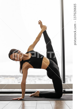 Young woman in black activewear practices an advanced kneeling side stretch against a large window with a city view 137026685