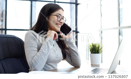 A young woman with glasses in the office is working on a laptop and talking on the phone A young woman with glasses in the office is working on a laptop and talking on the phone 137026807