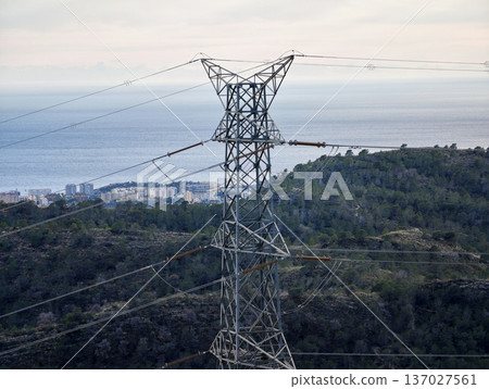 High voltage transmission on coast, Electrical transmission tower spanning landscape with distant town High voltage transmission on coast, Electrical transmission tower spanning landscape with distant town 137027561