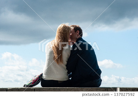 Man and woman sitting together on a stone wall, seen from behind, looking at dramatic clouds in blue sky. Concept of love, relationship, support, future and togetherness. 137027592