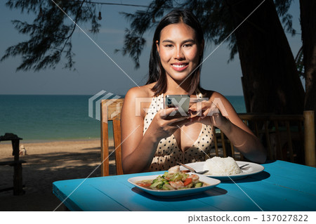Smiling Woman Using Smartphone While Dining at Beachside Cafe Smiling Woman Using Smartphone While Dining at Beachside Cafe 137027822