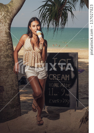 Smiling woman holding ice cream cone while standing on sandy beach near sign full length shot Smiling woman holding ice cream cone while standing on sandy beach near sign full length shot 137027823