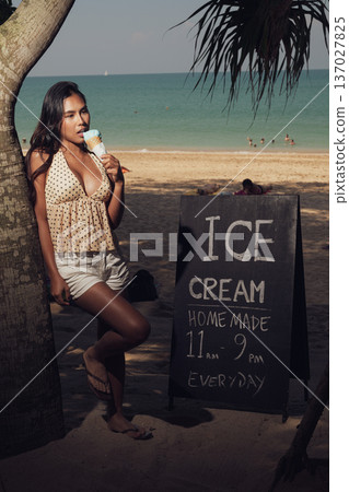 Smiling woman holding ice cream cone while standing on sandy beach near sign full length shot Smiling woman holding ice cream cone while standing on sandy beach near sign full length shot 137027825