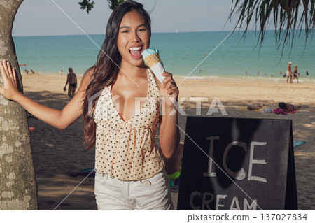 Smiling woman holding ice cream cone while standing on sandy beach near sign Smiling woman holding ice cream cone while standing on sandy beach near sign 137027834