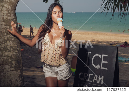 Smiling woman holding ice cream cone while standing on sandy beach near sign Smiling woman holding ice cream cone while standing on sandy beach near sign 137027835